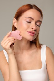 Photo of Young woman massaging her face with rose quartz gua sha tool on grey background