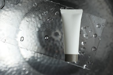 Photo of Moisturizing cream in tube on glass with water drops against metal background, top view. Space for text