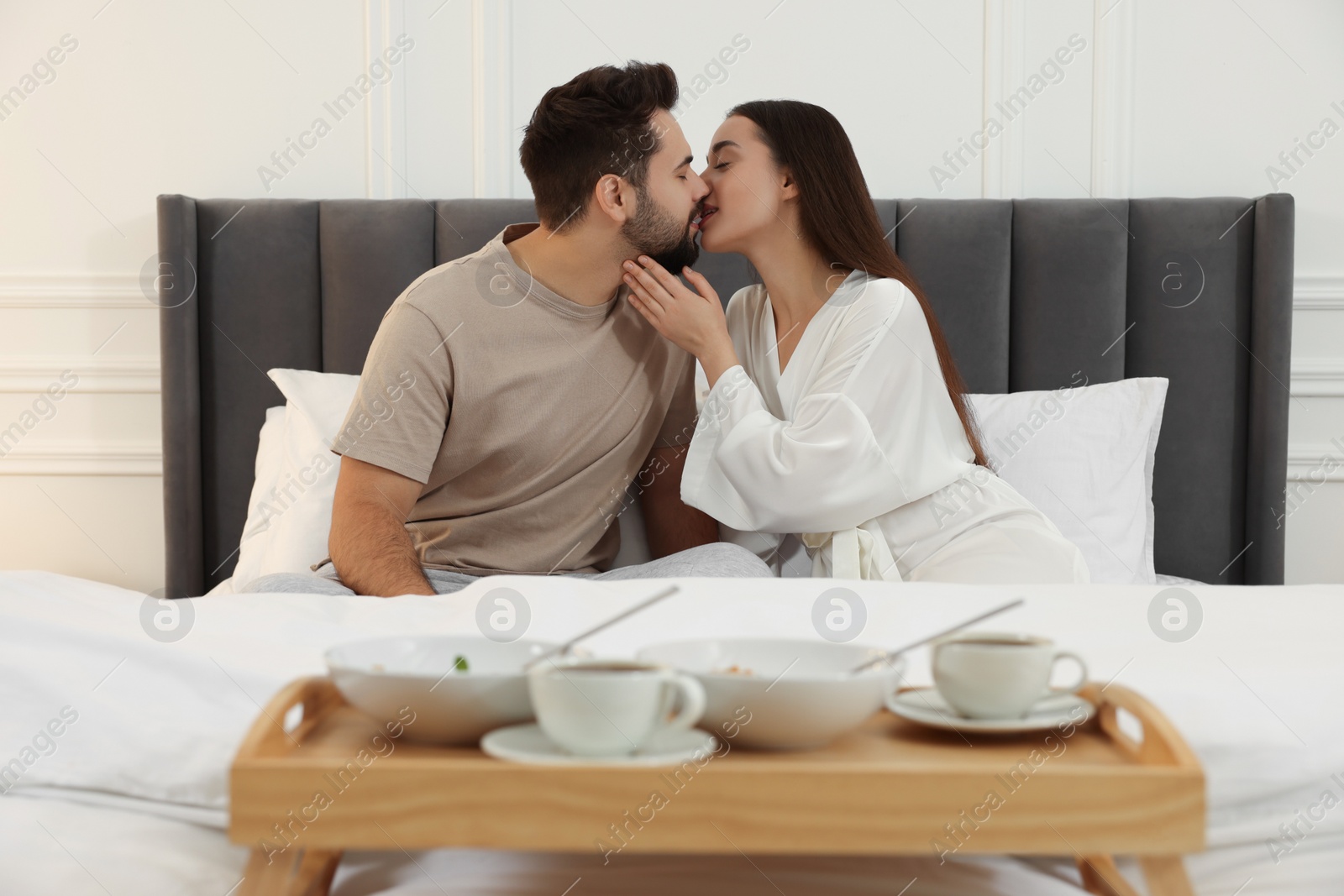 Photo of Happy couple kissing near wooden tray with breakfast on bed at home