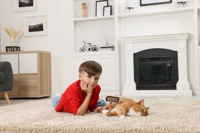 Photo of Little boy with pet brush and cute ginger cat on soft carpet at home