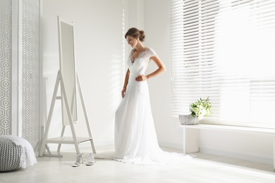 Young bride in beautiful wedding dress near mirror indoors