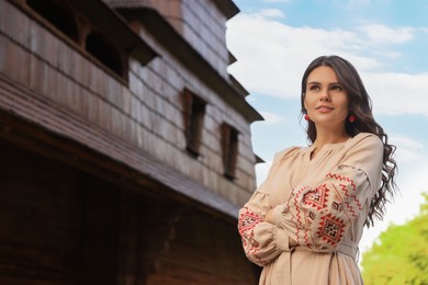 Photo of Beautiful woman wearing embroidered dress near old wooden church in village. Ukrainian national clothes