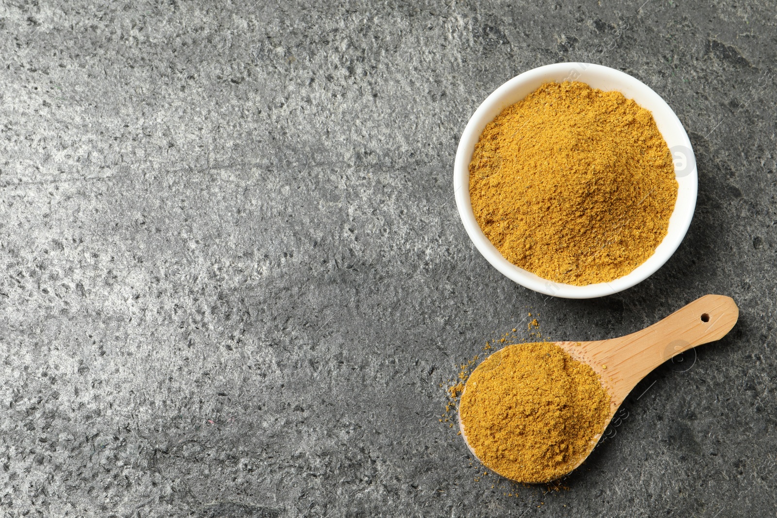 Photo of Spoon and bowl with dry curry powder on dark textured table, top view. Space for text