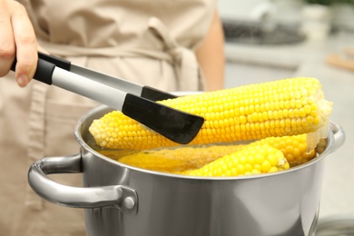 Photo of Woman taking boiled corn from pot with tongs in kitchen, closeup