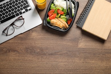 Photo of Container of tasty food, laptop, glasses and notebook on wooden table, flat lay with space for text. Business lunch