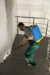 Pest control worker in uniform spraying pesticide on stairs indoors