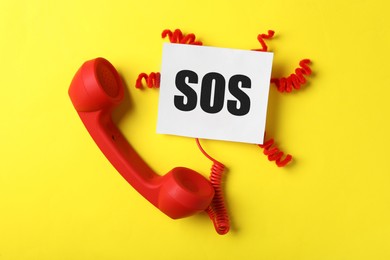 Image of Telephone handset and paper note with abbreviation SOS on yellow background, flat lay. Emergency call