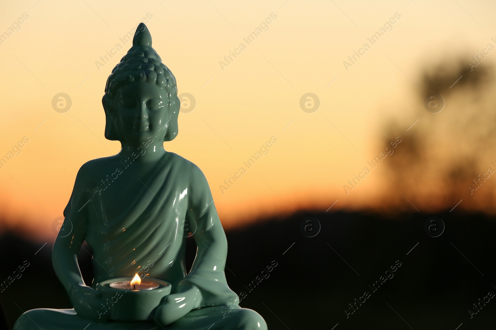 Photo of Decorative Buddha statue with burning candle outdoors at sunset. Space for text