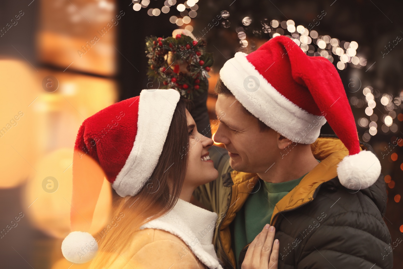 Photo of Happy couple in Santa hats standing under mistletoe wreath outdoors, bokeh effect