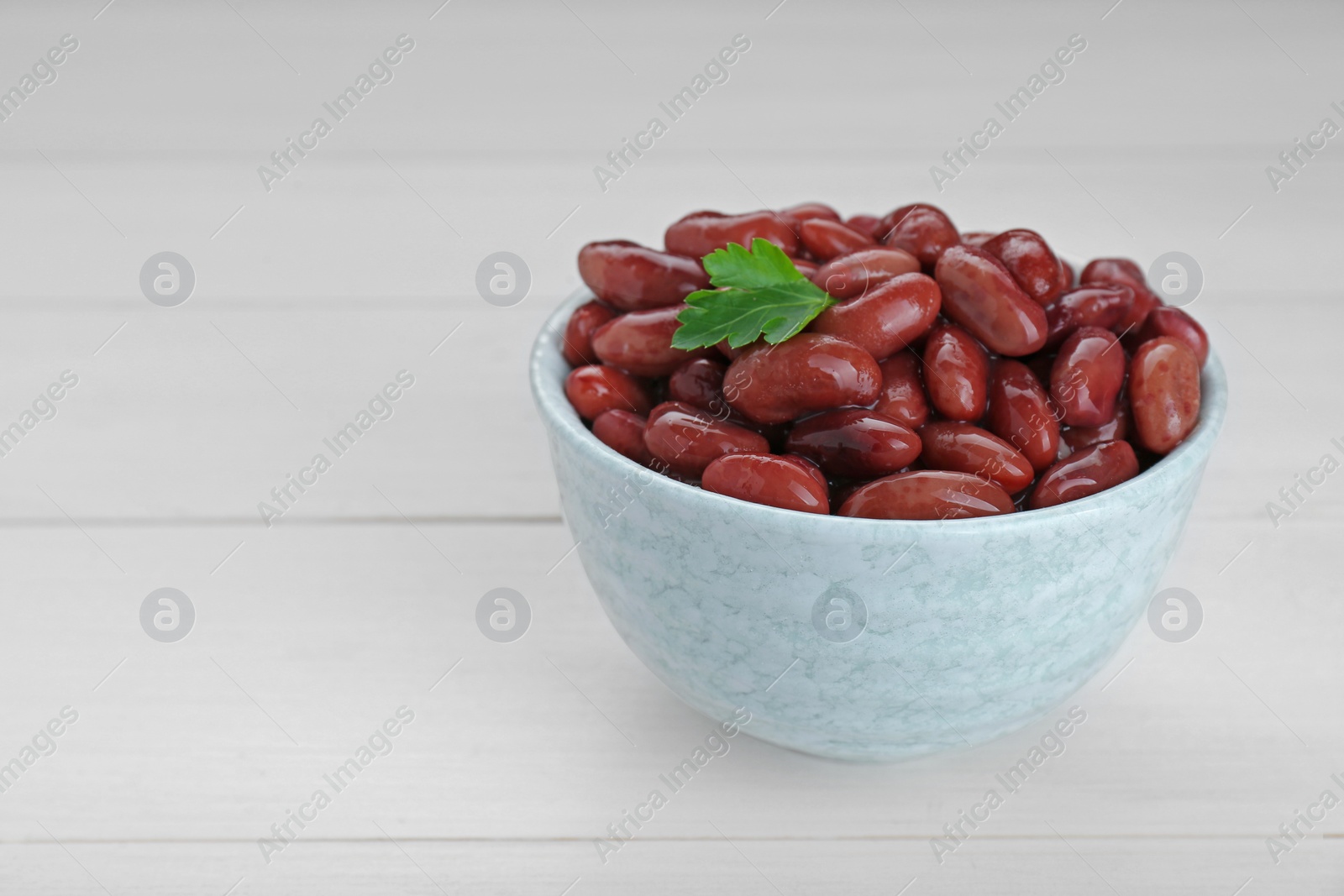 Photo of Bowl of canned kidney beans with parsley on white wooden table, closeup. Space for text