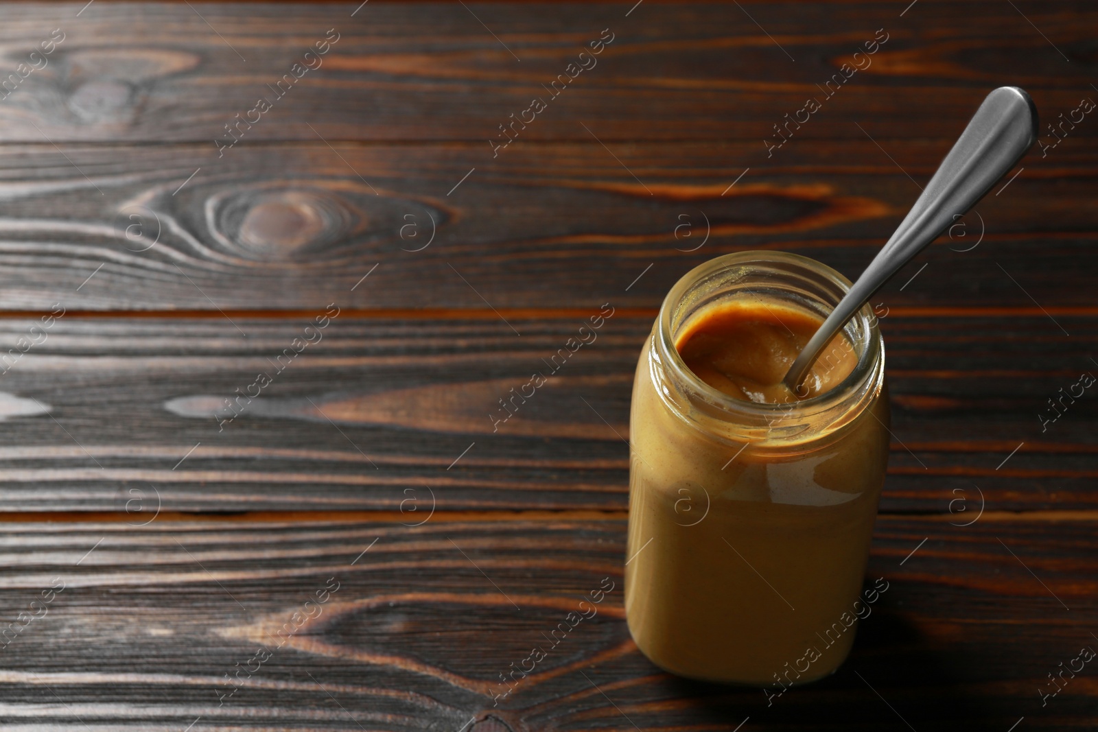 Photo of Spicy mustard and spoon in glass jar on wooden table. Space for text