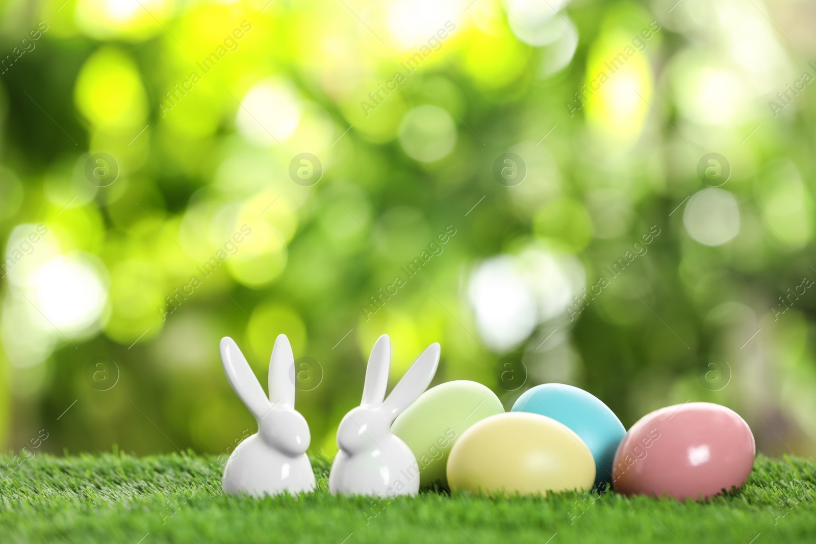 Photo of Ceramic Easter bunnies and dyed eggs on green grass against blurred background. Space for text