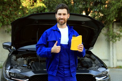 Photo of Smiling worker holding yellow container of motor oil and showing thumbs up near car outdoors