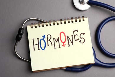 Photo of Notebook with word HORMONES and stethoscope on grey background, flat lay