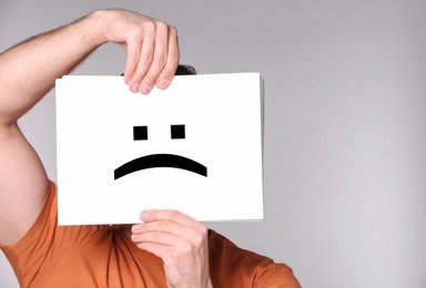 Image of Man hiding behind sheet of paper with sad face on grey background
