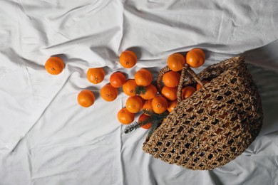 Photo of Stylish wicker bag with ripe tangerines on white bedsheet, above view