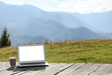 Photo of Modern laptop with blank screen and coffee cup on wooden surface in mountains, space for text. Working outdoors