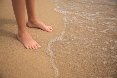 Photo of Little girl standing on sandy beach near sea, closeup. Space for text