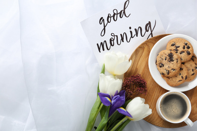 Photo of Delicious coffee, cookies, flowers and GOOD MORNING wish on white cloth, flat lay. Space for text