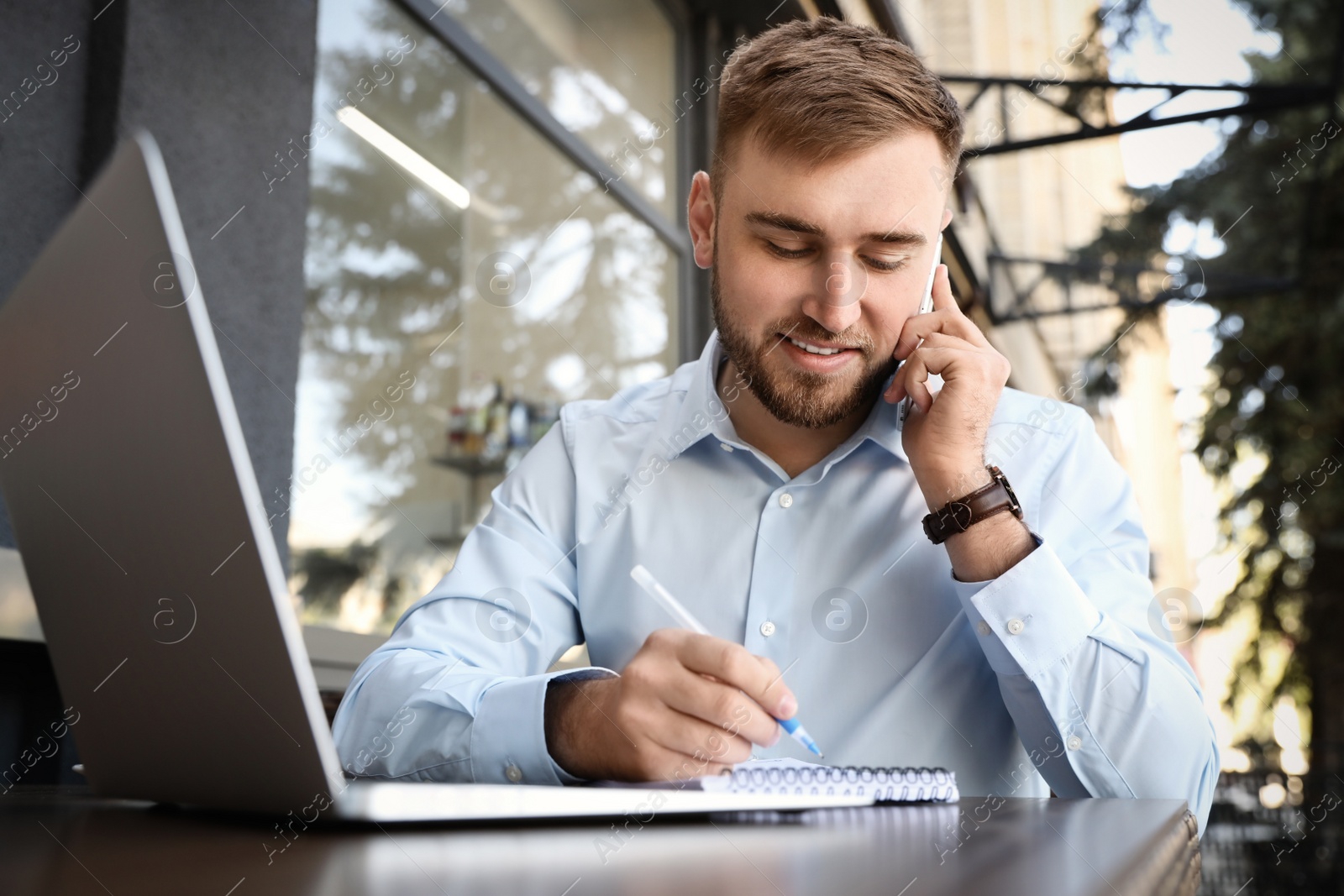 Image of Happy young man with laptop talking on phone at outdoor cafe