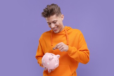 Happy man putting coins into piggy bank on purple background