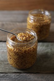 Photo of Jars and spoon of whole grain mustard on wooden table, closeup