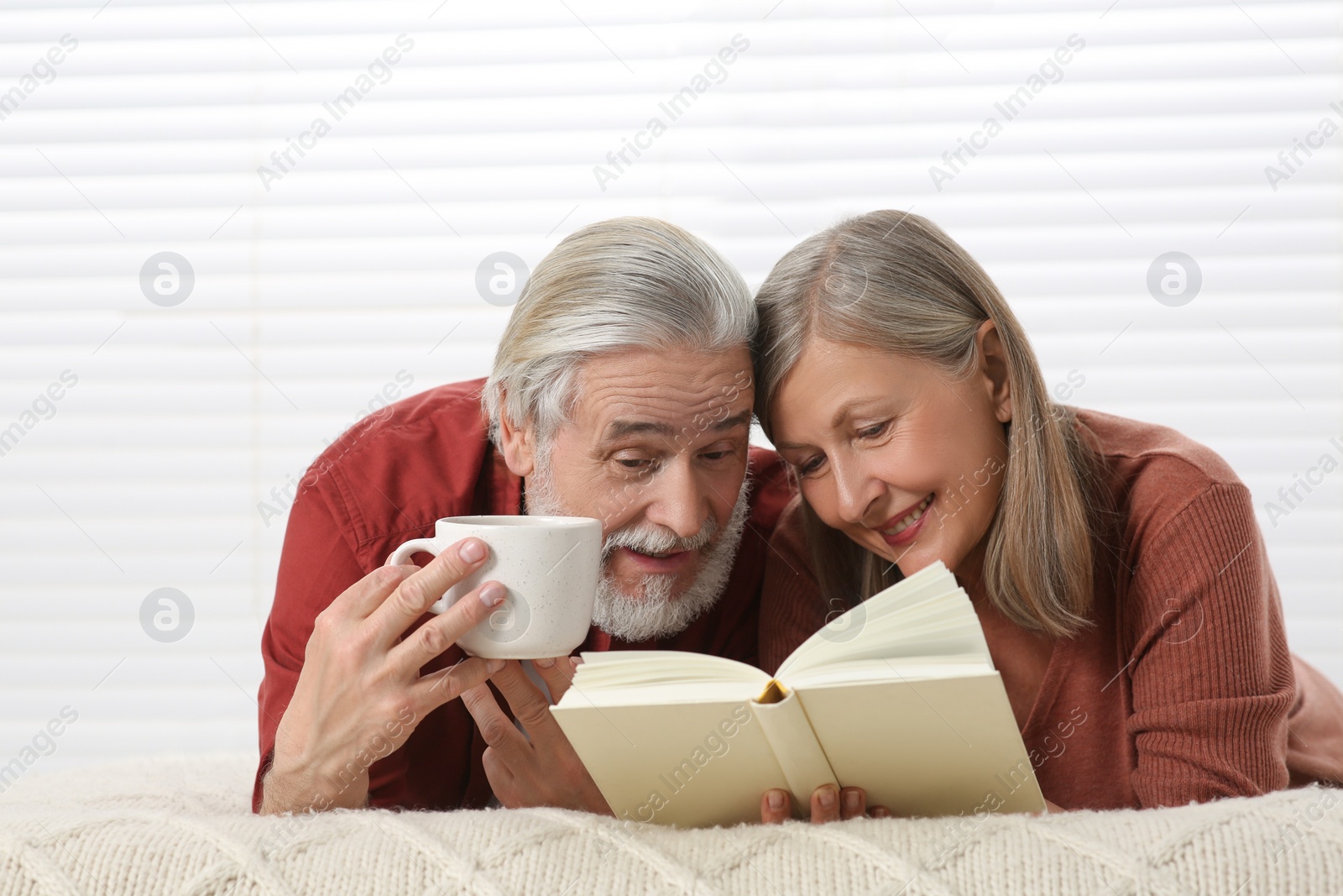 Photo of Senior man with cup of drink and his wife reading book on bed at home