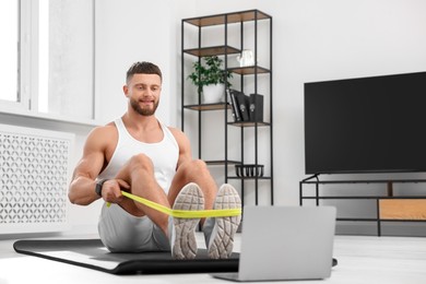 Muscular man doing exercise with elastic resistance band near laptop on mat at home