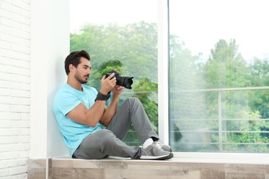 Young photographer with professional camera near window indoors. Space for text