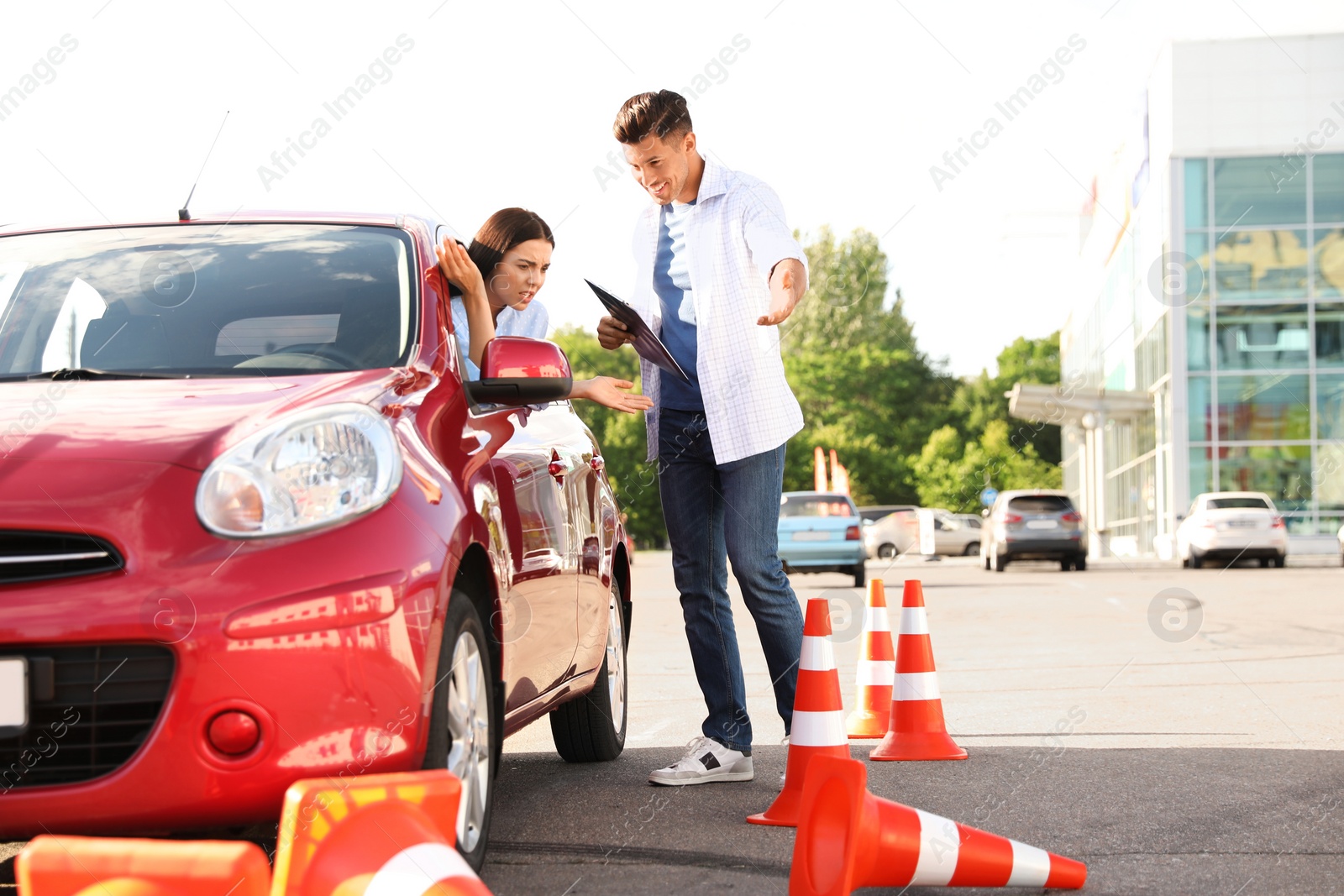 Photo of Stressed young woman in car near instructor and fallen traffic cones outdoors. Failed driving school exam