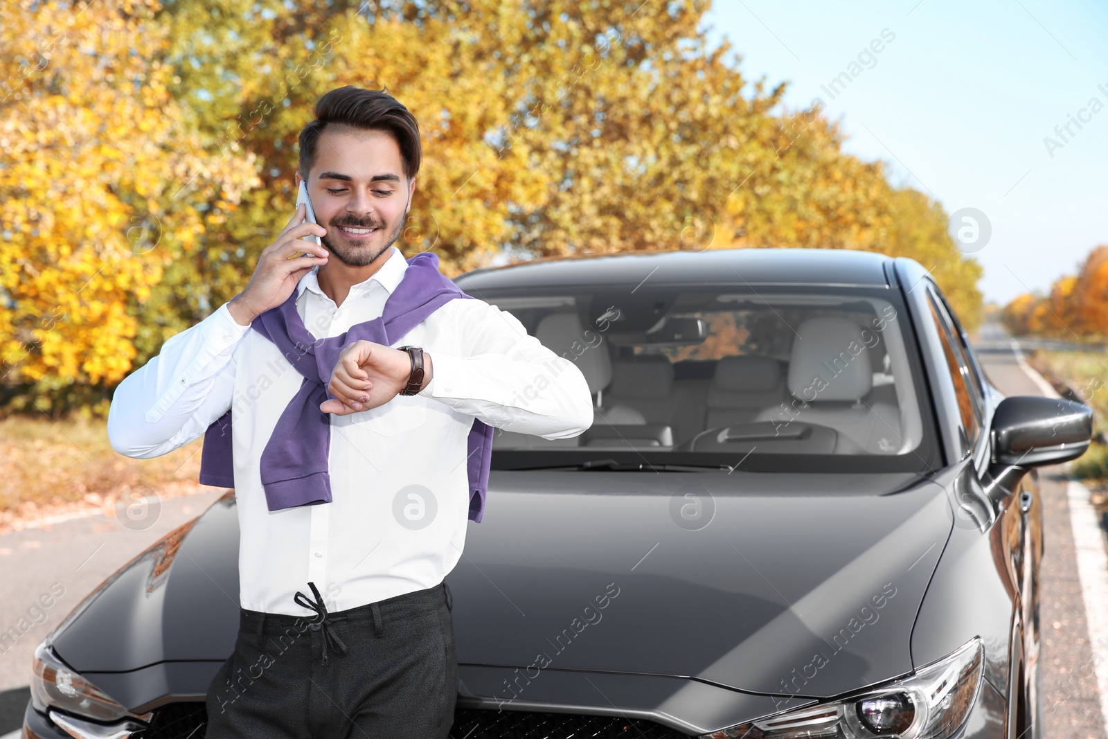 Photo of Young man checking time while talking on phone near modern car, outdoors