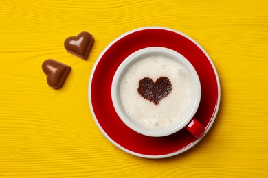Cup of aromatic coffee with heart shaped decoration and chocolate candies on yellow wooden table, flat lay
