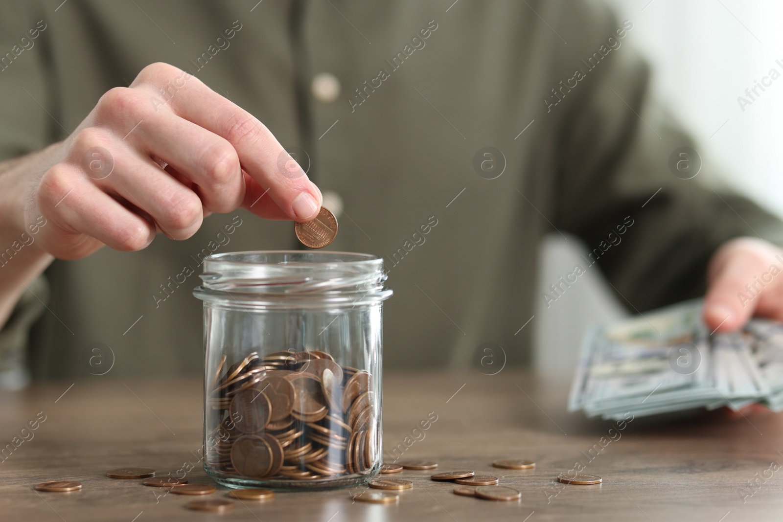 Photo of Financial savings. Man putting coin into glass jar at wooden table, closeup