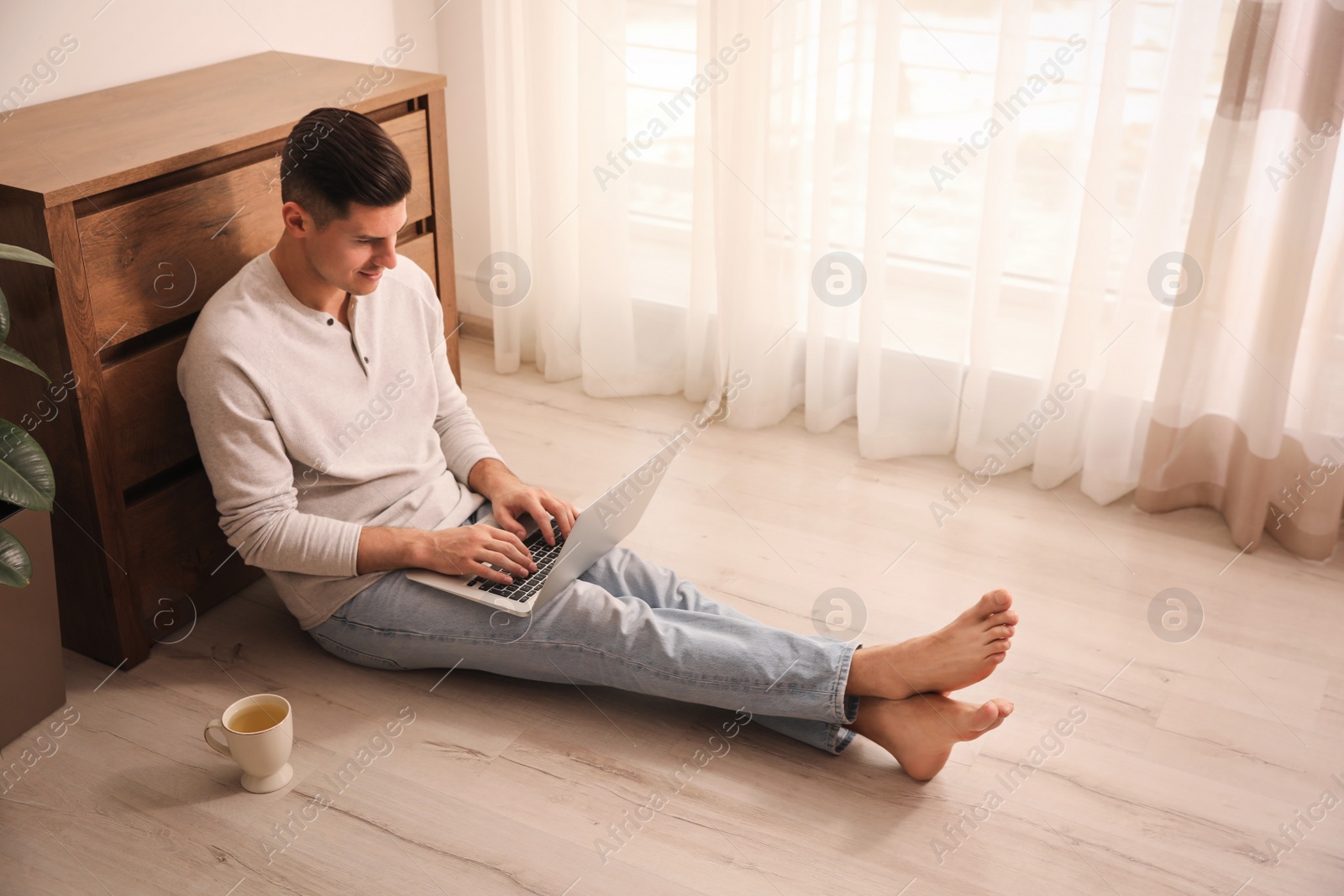 Photo of Man with cup of drink and laptop sitting on warm floor at home. Heating system