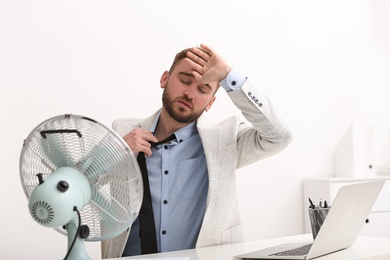 Photo of Man suffering from heat in front of fan at workplace