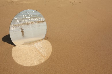 Photo of Round mirror reflecting sea on sandy beach, space for text
