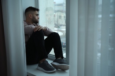 Sad man sitting on sill and looking at window indoors