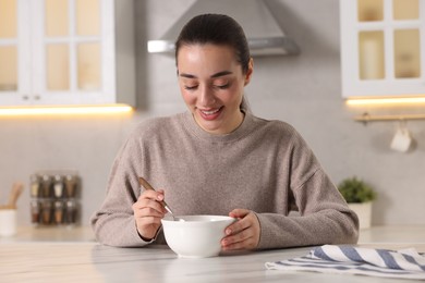 Smiling woman eating tasty soup at white marble table in kitchen