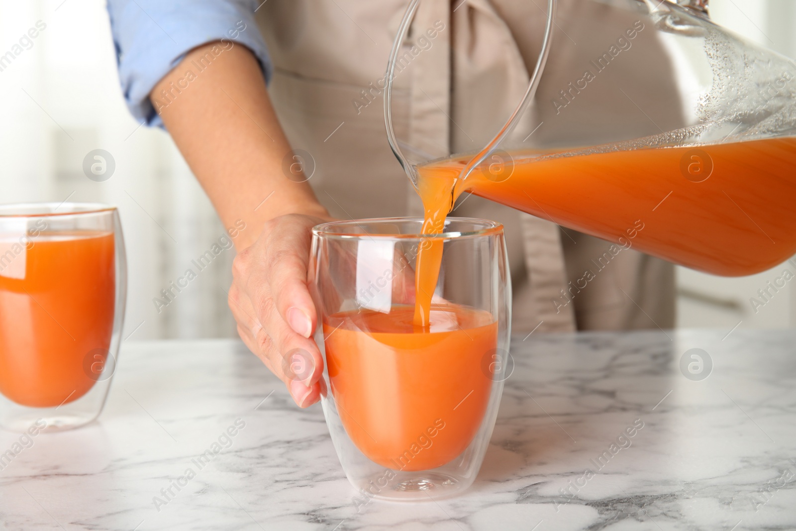 Photo of Woman pouring freshly made carrot juice into glass on white marble table in kitchen, closeup