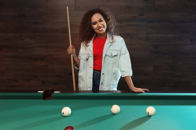 Photo of Young African-American woman with cue near billiard table indoors