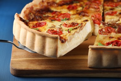 Photo of Taking piece of delicious homemade quiche with prosciutto, tomatoes and greens from board on blue table, closeup