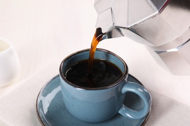 Pouring aromatic coffee from moka pot into cup at white table, closeup