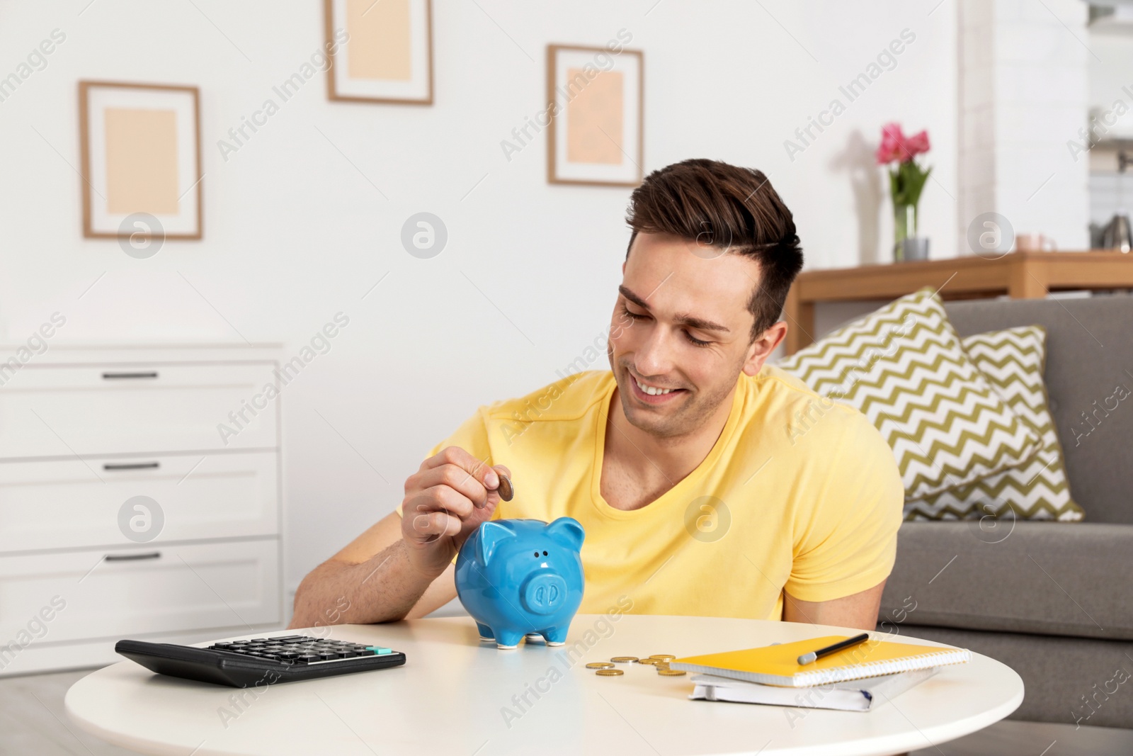 Photo of Happy man putting coin into piggy bank at table in living room. Saving money
