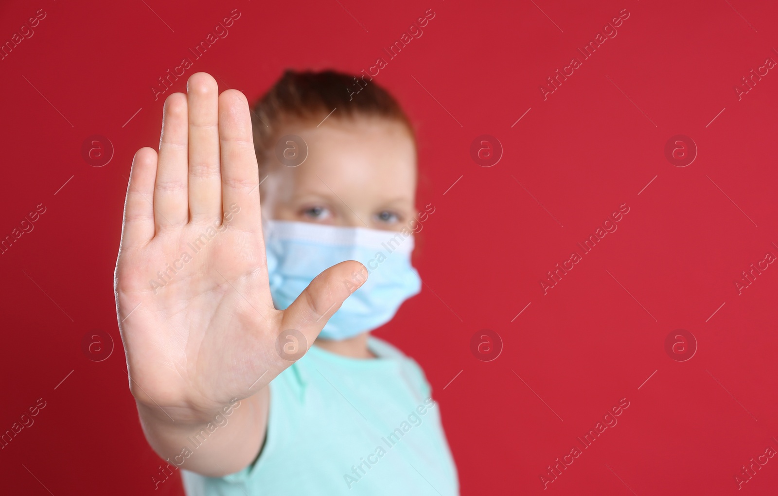 Photo of Little girl in protective mask showing stop gesture on red background, space for text. Prevent spreading of coronavirus