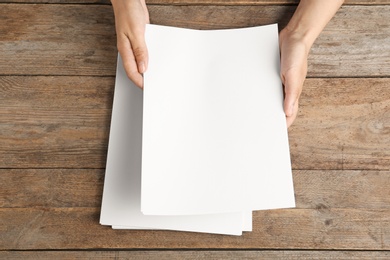 Photo of Woman holding blank paper sheets for brochure at wooden table, top view. Mock up