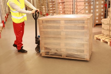 Photo of Worker moving wrapped wooden pallets with manual forklift in warehouse, closeup