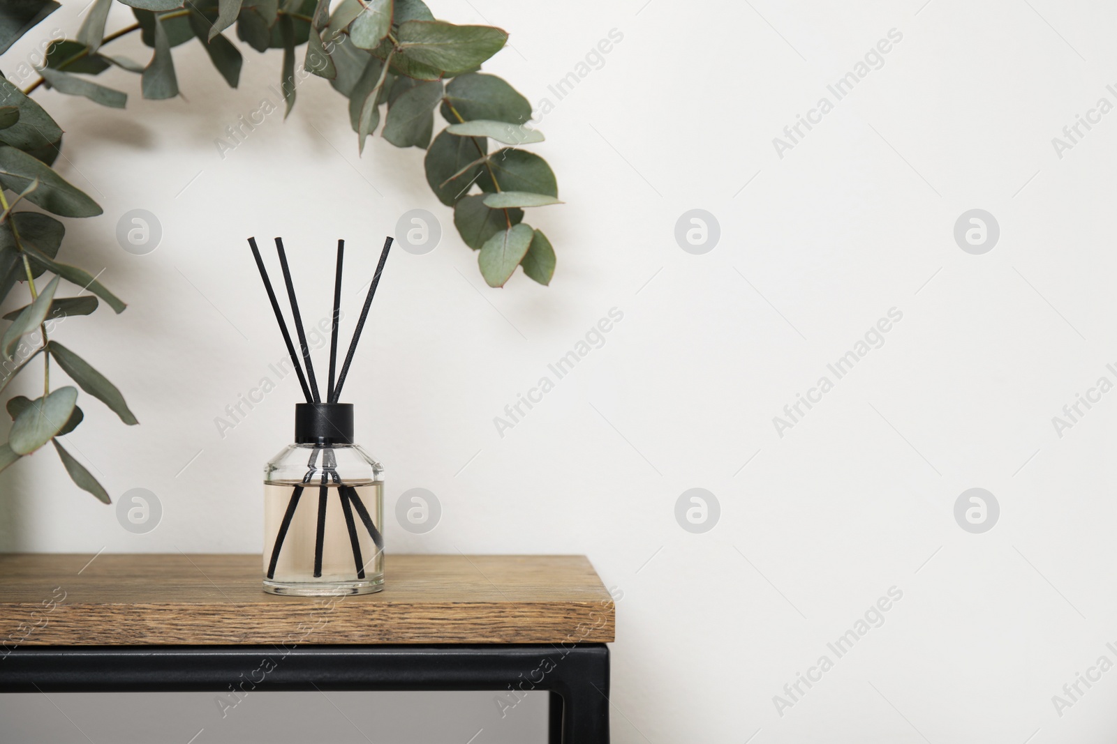 Photo of Reed diffuser and eucalyptus on wooden table near white wall. Space for text