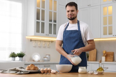 Making bread. Man pouring milk into bowl at wooden table in kitchen, space for text