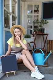 Photo of Young woman with blank chalkboard sitting on porch near home plant shop