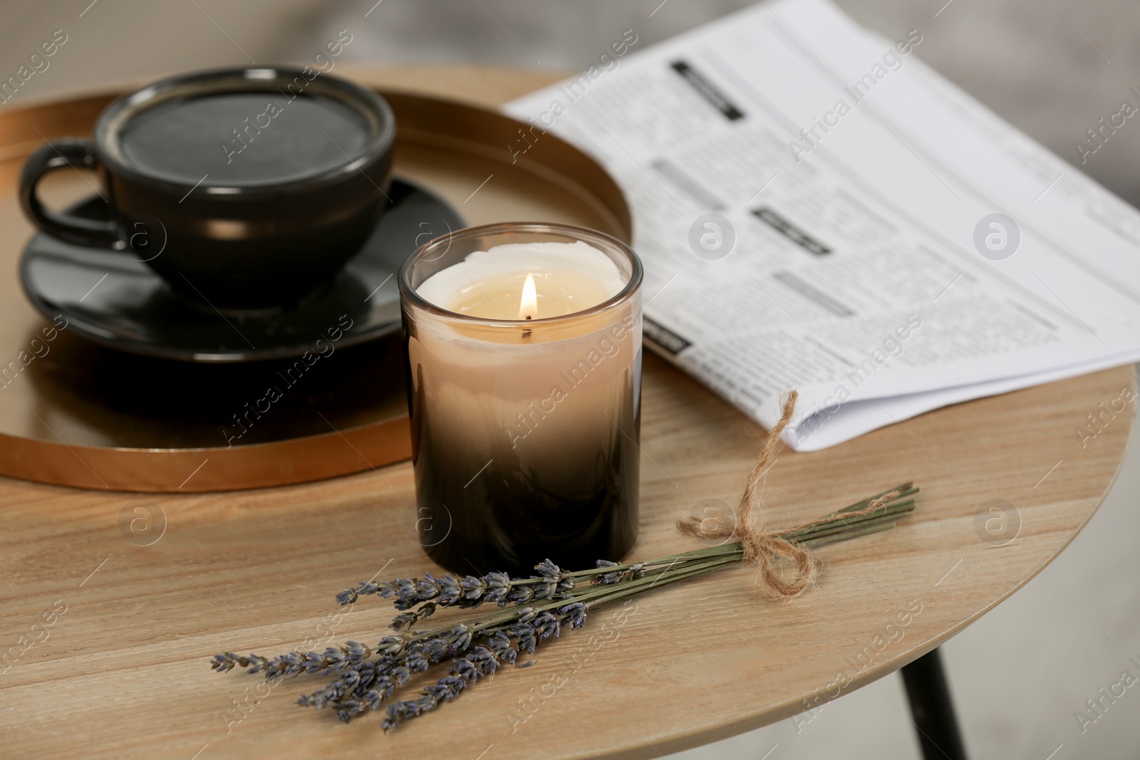 Photo of Beautiful candle, lavender, newspaper and cup of coffee on round wooden table indoors